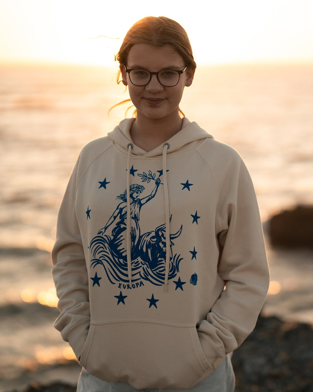Mädchen steht im Abendlicht am Strand mit dem Ankerherz Hoodie EUROPA ONE LOVE Ivory Garment Dye, blauer Siebdruck Europa auf Zeus-Stier mit zwölf Sternen von Dani Garreton, Unisex Frontansicht
